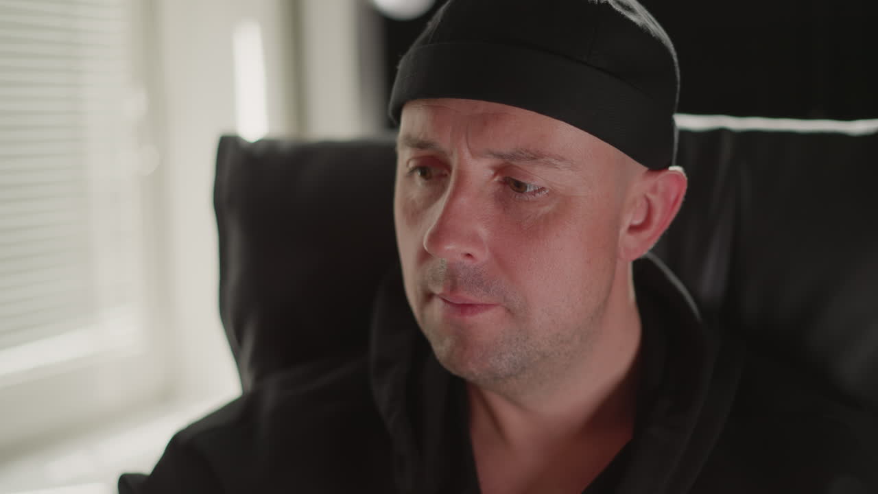 Close-up of a man wearing a black cap and black shirt,sitting on a black chair with a concentrated expression. He appears to be intently looking at something out of frame, conveying a sense engagement