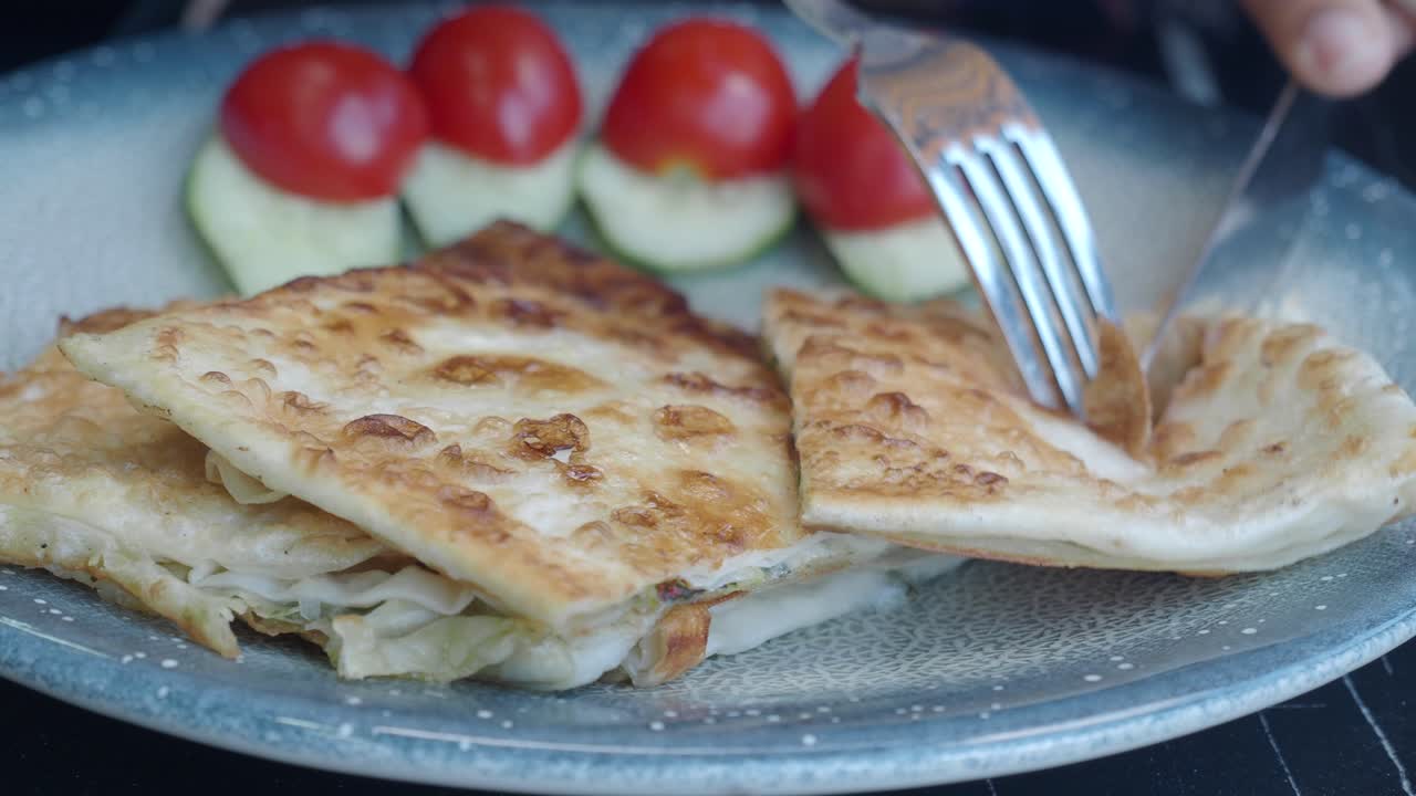 Close-up of a delicious Gozleme dish being cut with a fork and knife. Served with tomatoes and cucumber.