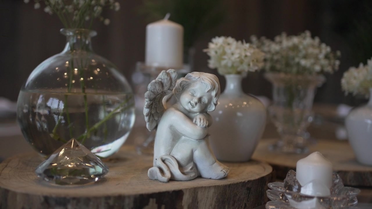 Una statuetta di angelo è posizionata su una lastra di legno, circondata da vasi di vetro e bianchi con fiori delicati, e una piccola candela in un portacandele di cristallo.