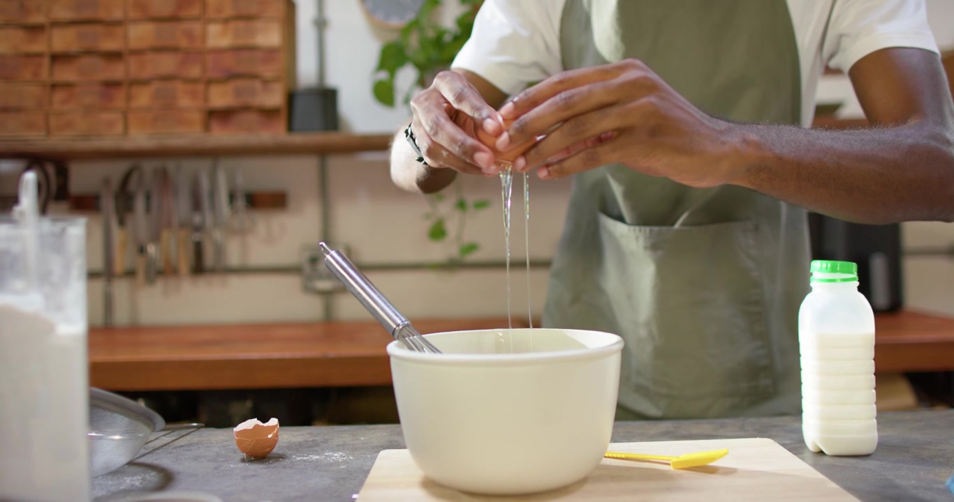 African man in green apron preparing batter by cracking eggs into mixing bowl at kitchen counter