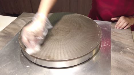 worker cleaning a round crepe maker in a professional kitchen, hand in glove performing a circular wipe motion.