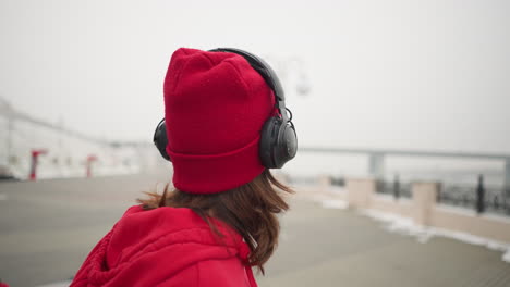 woman in red beanie and hoodie wearing headphones, looking contemplative with mouth slightly open, during winter, background features decorative fence, blurred atmospheric fog, and an urban setting