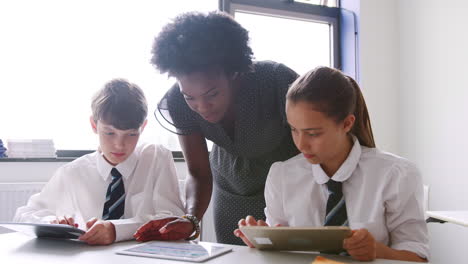 Female-High-School-Teacher-At-Table-With-Students-Wearing-Uniform-Using-Digital-Tablets-In-Lesson