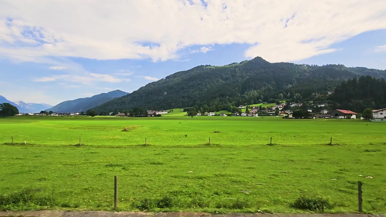 澄み切った青い夏の空の下、緑の谷、シャレーの屋根、並木、そして暗い山脈が広がる、走行中の列車から見たオーストリアアルプスの美しい風景