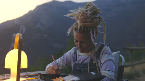 Hippy-girl-with-blond-dreadlocks,-deep-in-thought-draws-or-writes-outdoors