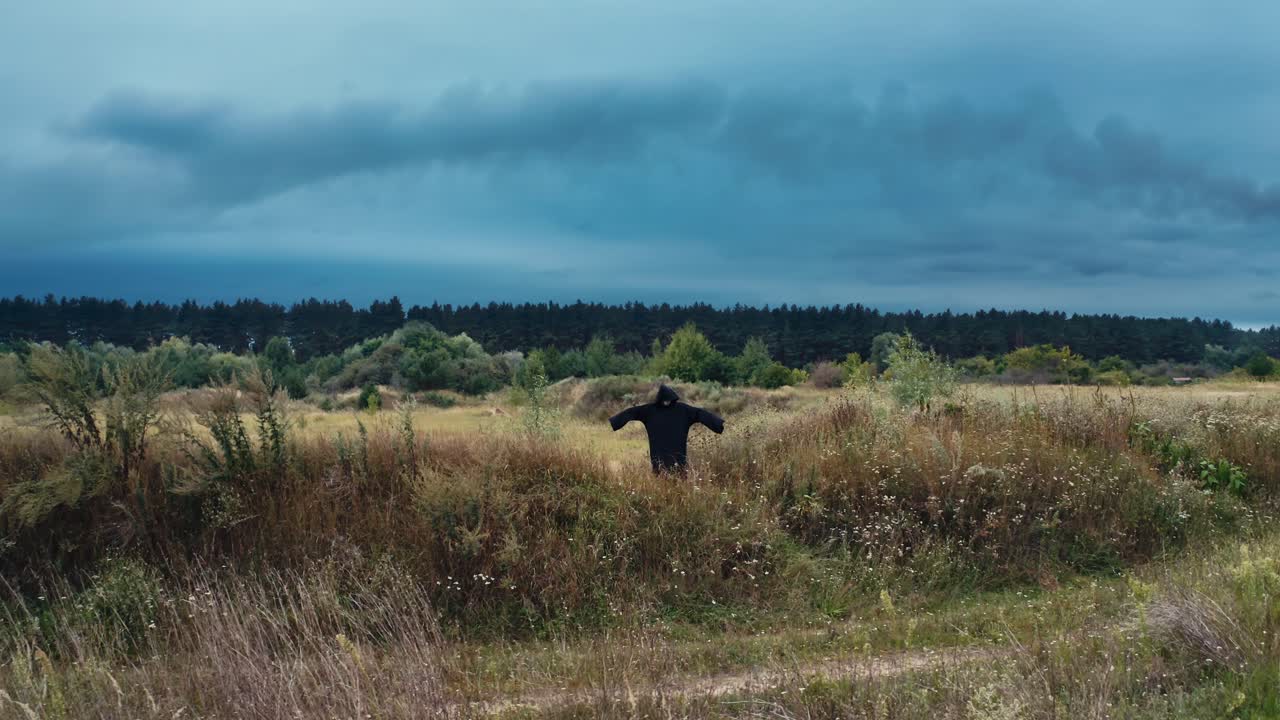 Dark scary figure on the field. Black death in mantle standing with outstretched arms among summer field under evening sky.
