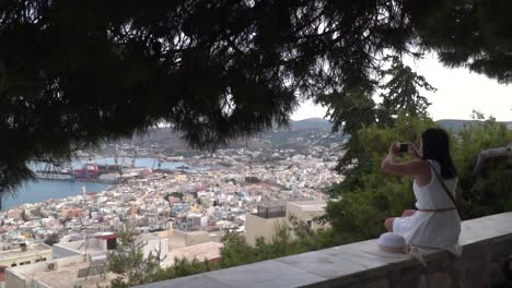 chica con vestido blanco de verano tomando fotos de la ciudad con un smartphone desde el punto de vista mientras se sienta en la pared bajo un árbol