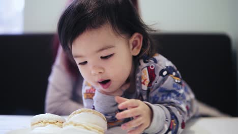 an adorable cute asian toddler boy enjoys eating macaroons with his mother sitting beside him.