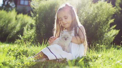 small girl sitting on the green grass on a sunny summer day and holding white kitty cat