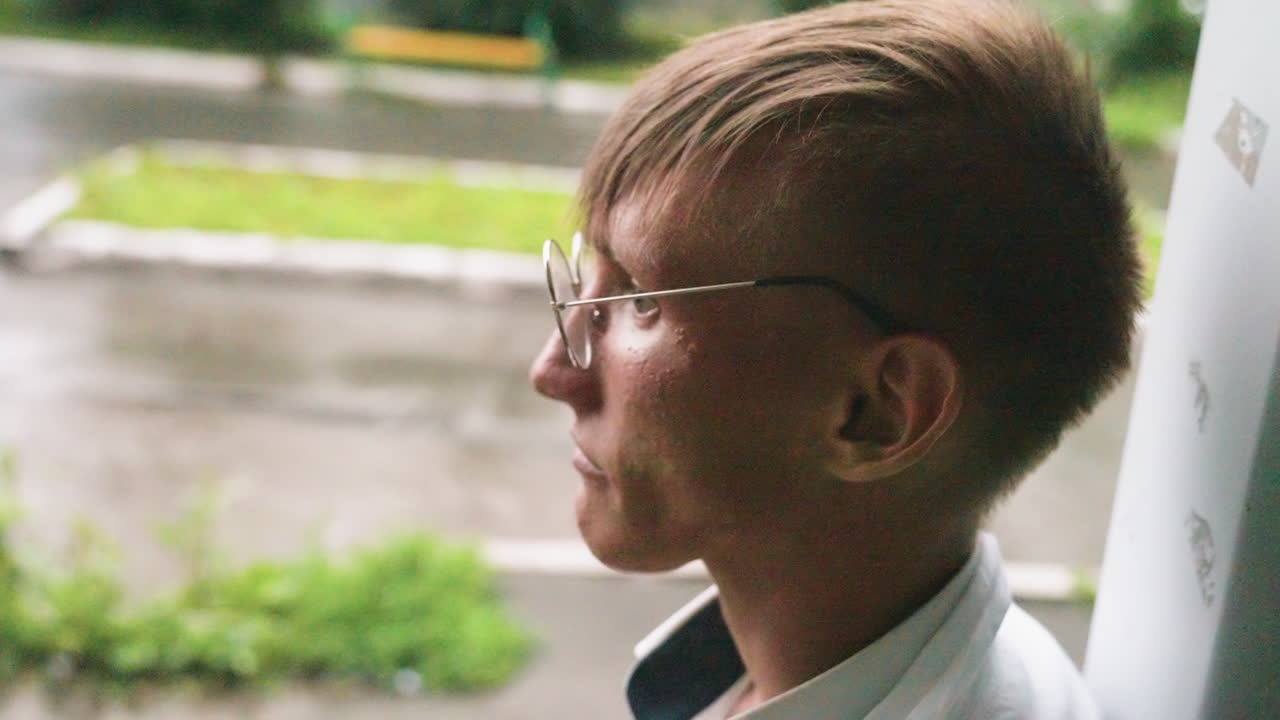 Botany student in white coat leans back against wall sipping juice during outdoor break, relaxing in calm urban environment with greenery and blurred pavement in background