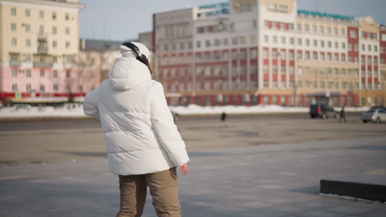 Back view of creative woman in white winter coat, beanie and headphones singing and dancing joyfully on paved urban plaza with passing cars and pedestrians, snowbanks under bright winter daylight