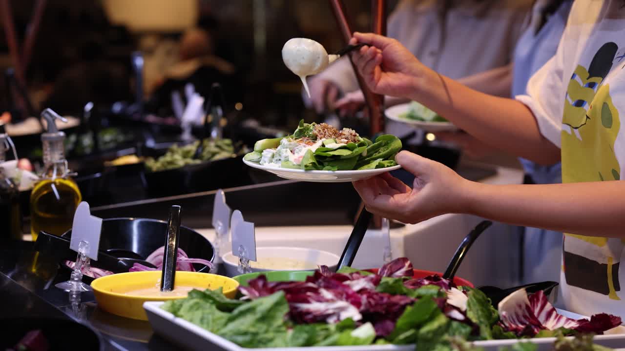 Person adds creamy dressing to mixed greens salad at self-serve salad bar under warm lighting