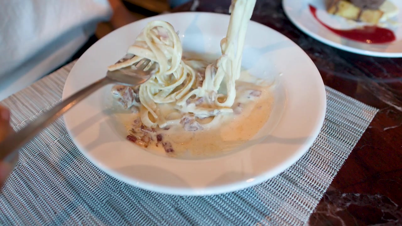 Primo piano di una persona che usa le forchette per arrotolare cremose fettuccine alla carbonara in un ristorante.
