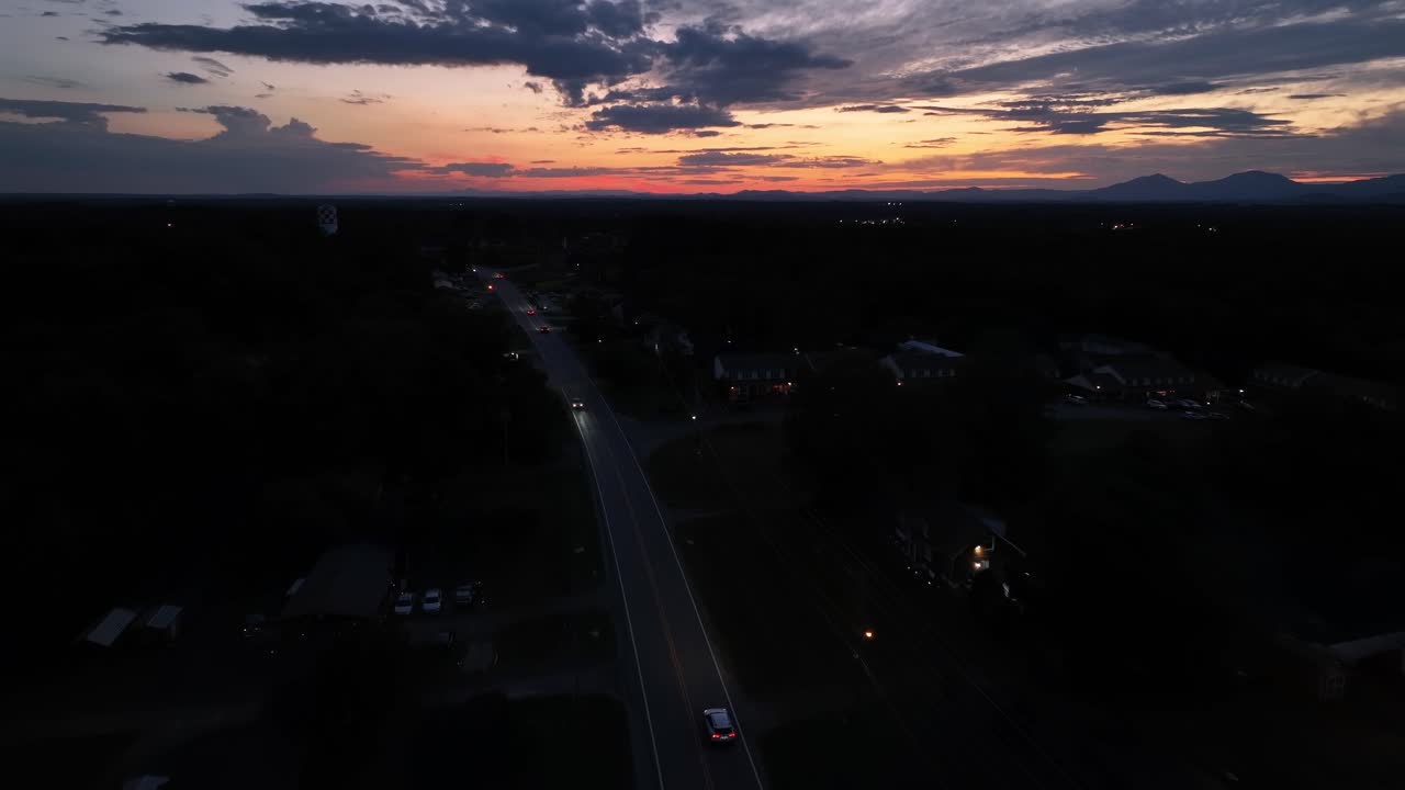 Aerial tracking shot of cars on suburb road between houses in American neighborhood at golden sunset. Dark Suburb settlement and bright sky at horizon. Wide shot