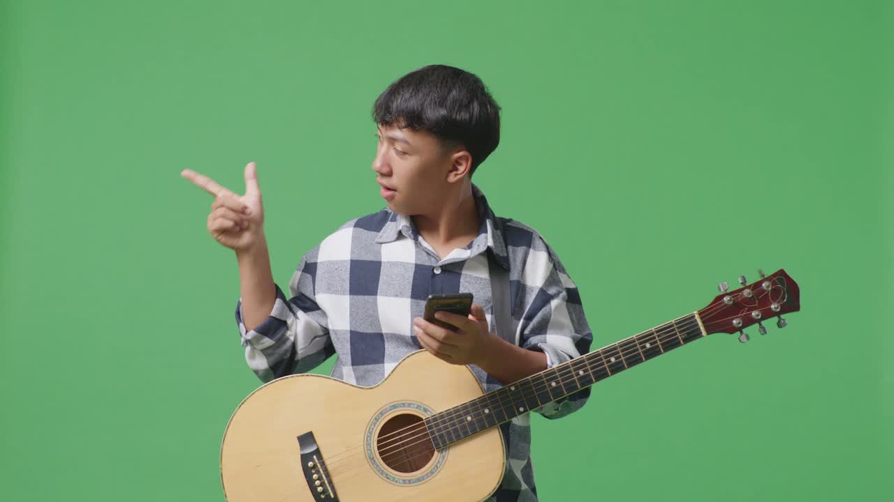 스마트폰 을 사용 하고 초록색 화면 배경 에 서 있는 동안  을 가리키는 기타 를 들고 있는 아시아 청소년