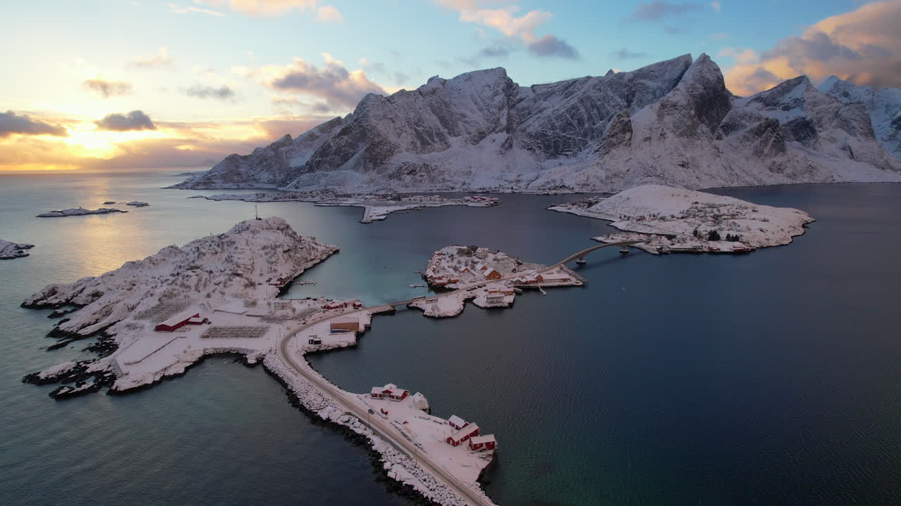 Cinematic 4K drone shot of Sakrisoy village. Iconic yellow rorbuer cabins and E10 bridge against snow capped Reinefjorden mountains at Arctic sunset. Rare winter light over the Norwegian fjords