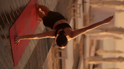 vertical video: rear view of a brunette girl in a black sports summer uniform doing a plank on one arm and reaching up with the other. yoga and sports class on a sunny beach with umbrellas in summer