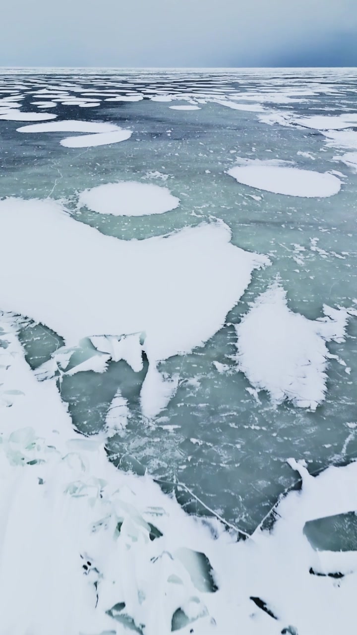 Drone glides over a frozen Baltic bay where a long snow strip cuts through dark slush and weak ice, creating a stark winter hazard scene with strong negative space, vertical.
