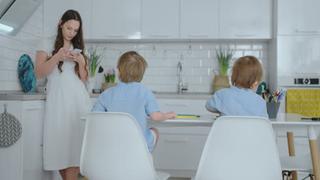 Dos-Hijos-Con-Camisas-Azules-Sentados-En-La-Mesa-Y-Dibujando-Lápices-Sobre-Papel-Sentados-En-Una-Mesa-Blanca,-Y-Mi-Madre-Escribe-Un-Mensaje-De-Texto-En-El-Smartphone.-Mamá-Empresario