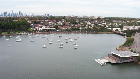 Aerial-drone-shot-flying-past-UTS-boat-house-with-Sydney-city-skyline-in-the-distance,-Australia