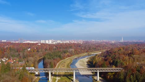 Clip-De-Viaje-Encantador---Volando-En-Drones-En-La-Temporada-De-Otoño-Sobre-El-Río-Isar,-Un-Tren-Pasa-Por-Un-Puente-Con-La-Vista-A-La-Popular-Ciudad-Turística-De-Munich-En-El-Sur-De-Alemania-Bajo-Un-Cielo-Azul