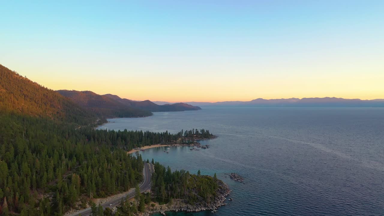 Aerial Drone View of Beautiful Lake Taho Water and Forest at Sunrise with a Road or Highway Going Through a Pine Trees and Mountains in the Background over a Beach