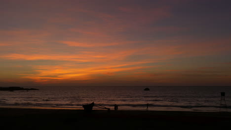 Silhouettes-of-People-with-Traditional-Fishing-Boat-on-Beach-at-Sunset,-Goa,-India