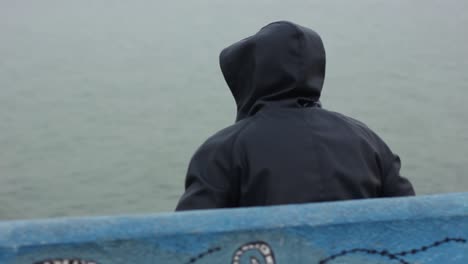 Un-Hombre-Encapuchado-Vestido-Con-Jeans-Azules-Sentado-En-Un-Banco-De-Concreto-Lleno-De-Graffiti-Azul-Mirando-El-Mar-Con-Una-Mirada-Incierta