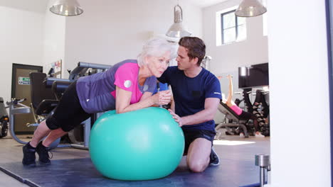 senior woman exercising on swiss ball being encouraged by personal trainer in gym