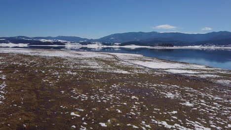 Drone-flies-over-a-lake-with-a-reflection-of-a-mountain-and-sky-in-winter