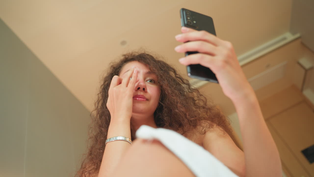 Selfie allo specchio di una camera d'hotel che mostra una donna in asciugamano che controlla il telefono e si tocca il viso, ritratto spontaneo dal basso, calda illuminazione ambientale, soffitto e porta dell'hotel sullo sfondo, stato d'animo disinvolto e sicuro.