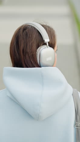 Back view of young girl in sweater and headphones walking, looking towards blurred iron rail in background, calm, relaxed outdoor scene with focus on her and surrounding environment