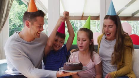 portrait of caucasian girl in party hat blowing candles on birthday cake while family smiling and wa