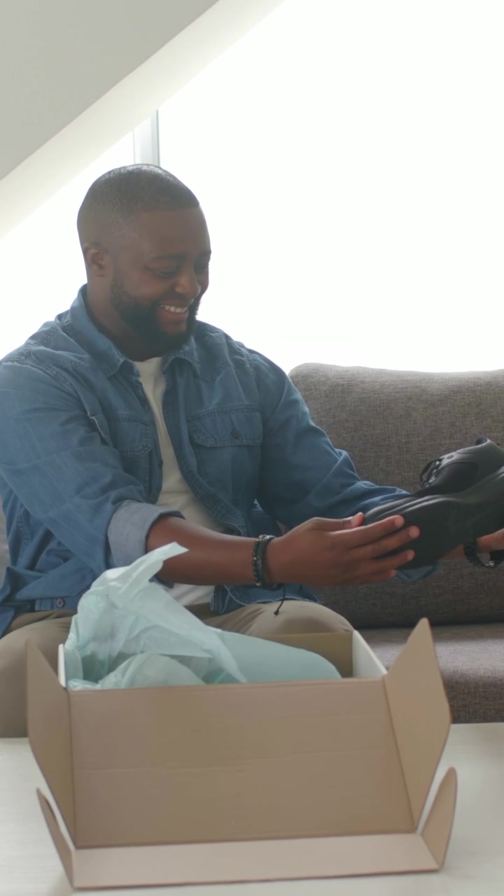 Vertical video: Reaching into shoebox, man in denim checking black shoe on couch for fit