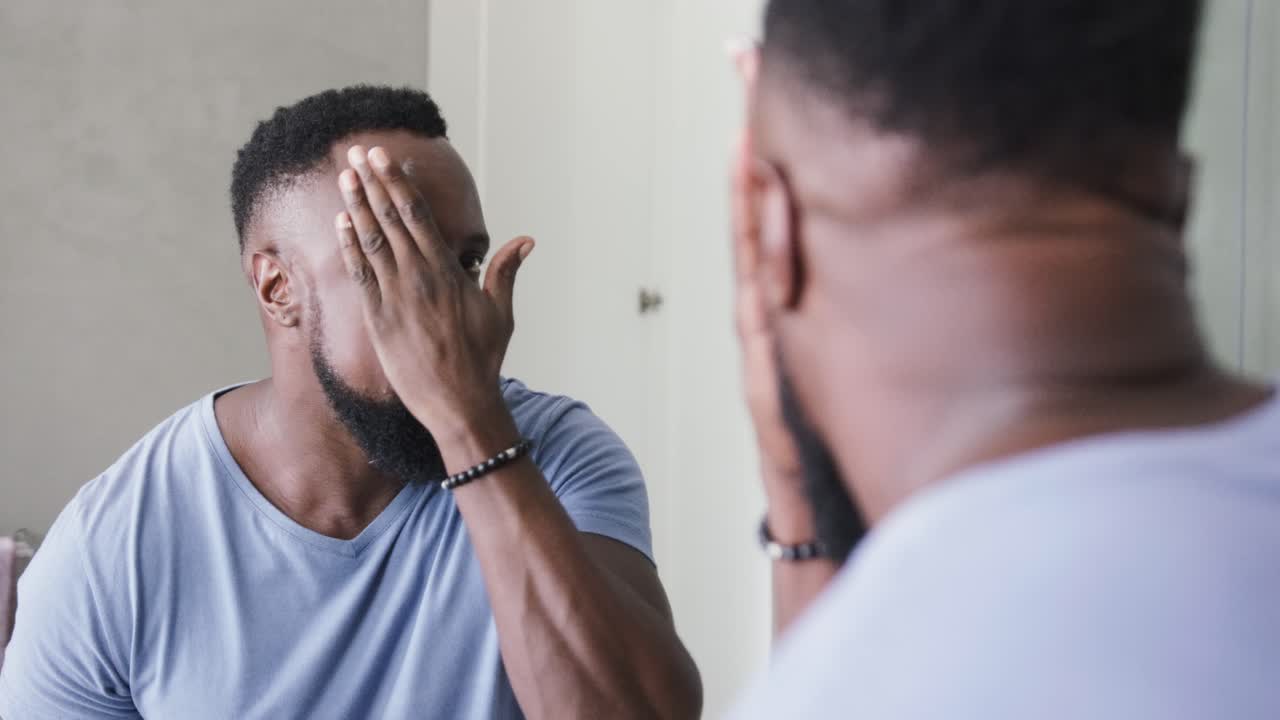 Adult African man noticing forehead dab, leaning to bathroom mirror, spreading cream for skincare