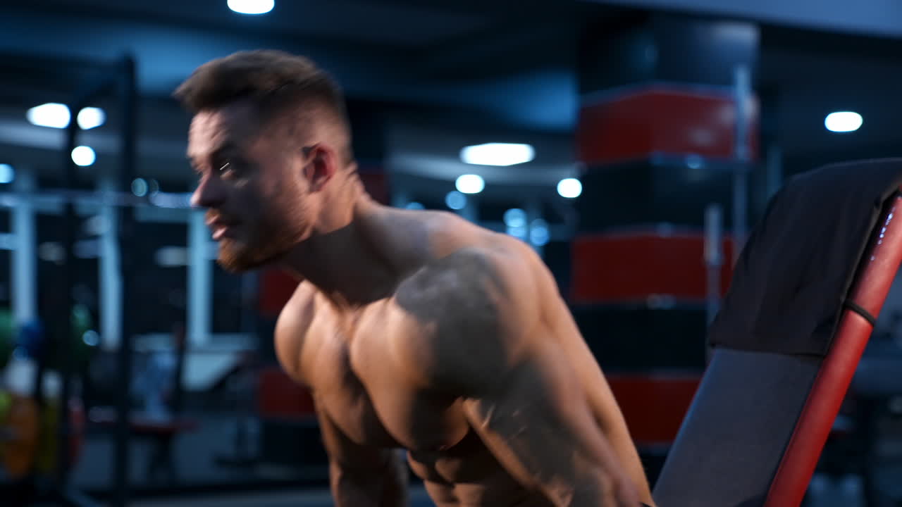 Shirtless bodybuilder in gym. Young guy with muscular body is lifting dumbbells. Sportsman practicing at sports center while sitting. Hard strength workout. Profile view.