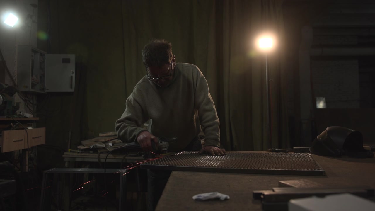 Adult man wearing glasses grinds metal sheet at workbench inside dim industrial workshop, with small sparks, warm backlight, deep shadows, rough textures, tools, cabinets, curtains, helmet, and