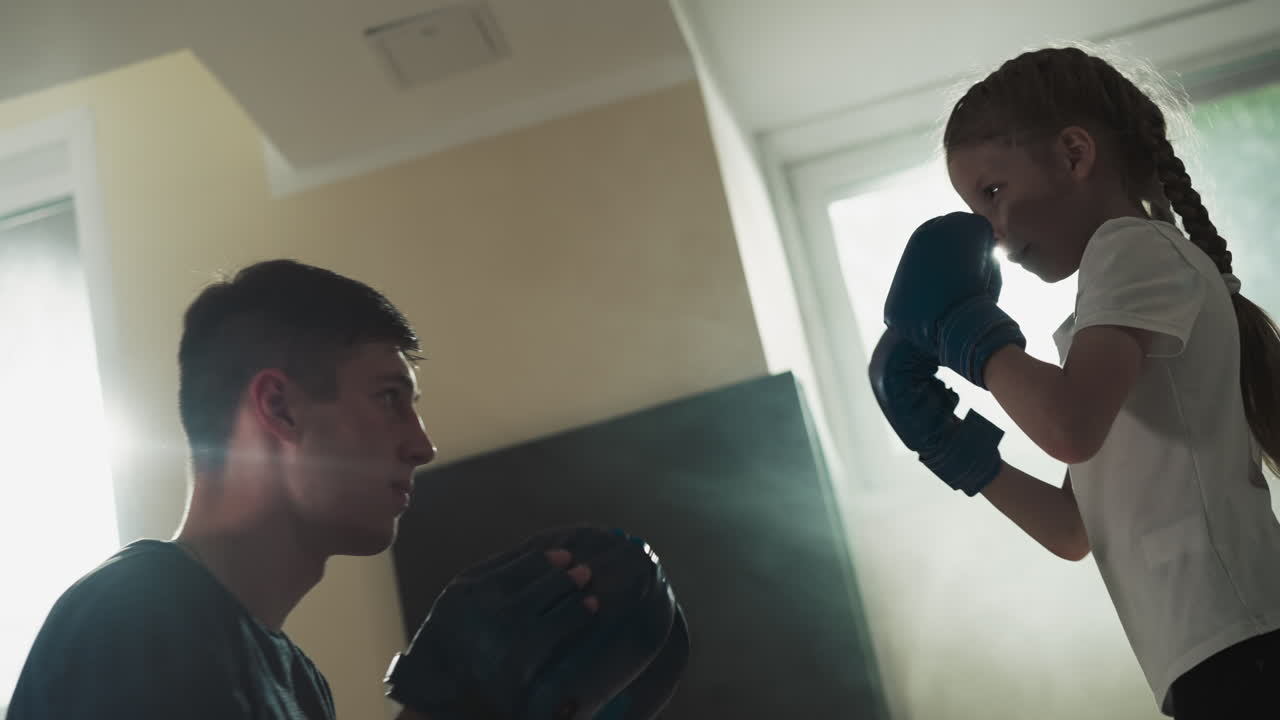 강사와 아이가 체육관에서 스파링을 하고, 조심스러운 트레이너가 장갑을 끼고 스포츠 클럽에서 소녀를 훈련시니다.