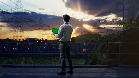 full body back view of an asian male professional worker standing with green screen tablet near high voltage tower, industrial facility