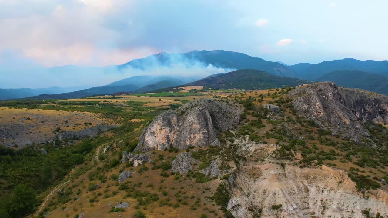 Reverse aerial starts tight on metal cross near Ilindentsi, then backs away to frame hill crest as grey smoke columns from active Pirin Mountain forest fire billow under shimmering summer sky