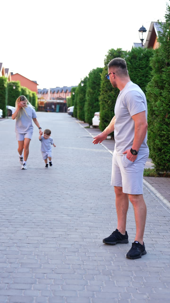 Family walk in quiet neighborhood. A man watches as a child plays on a quiet street lined with hedges, while a woman adjusts her child's outfit nearby