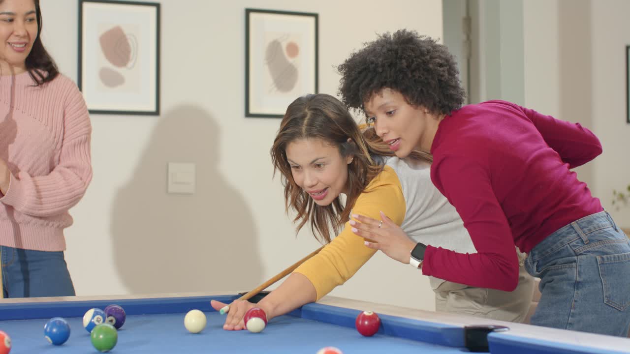 African American woman guiding Asian woman aligning cue and striking ball on pool table at home