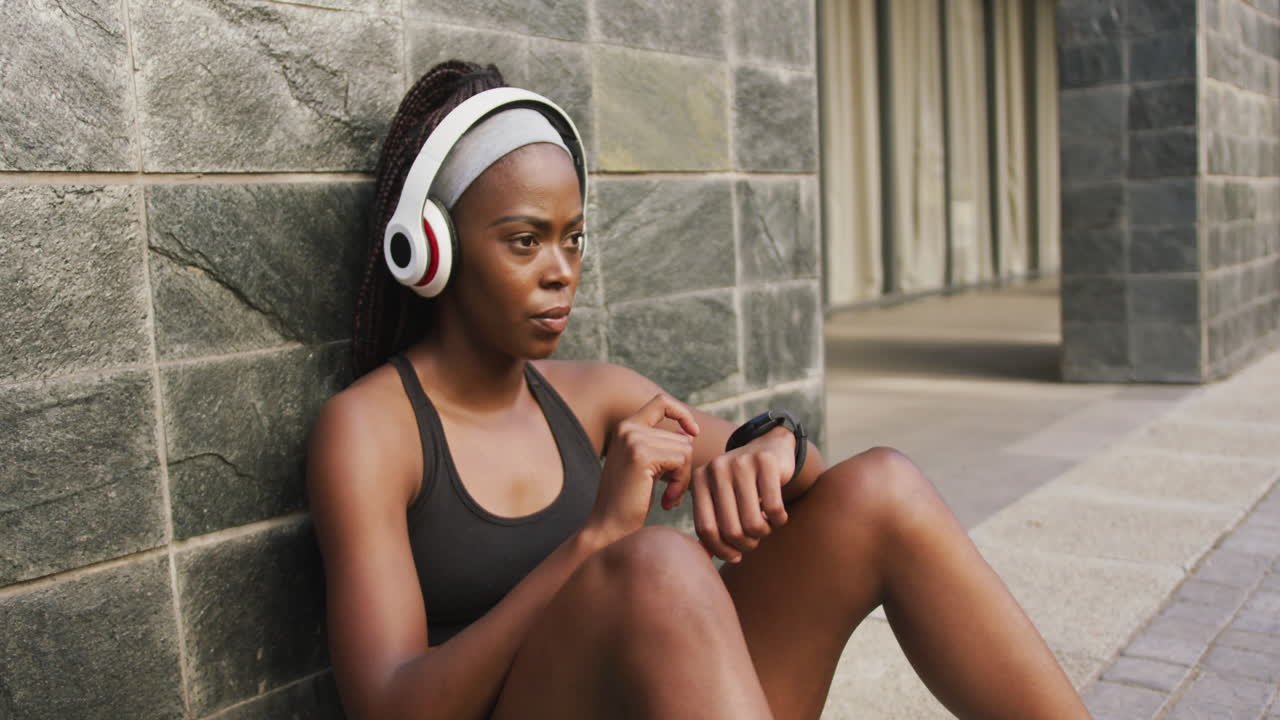 mujer afroamericana haciendo ejercicio al aire libre usando auriculares inalámbricos usando un reloj inteligente en la ciudad
