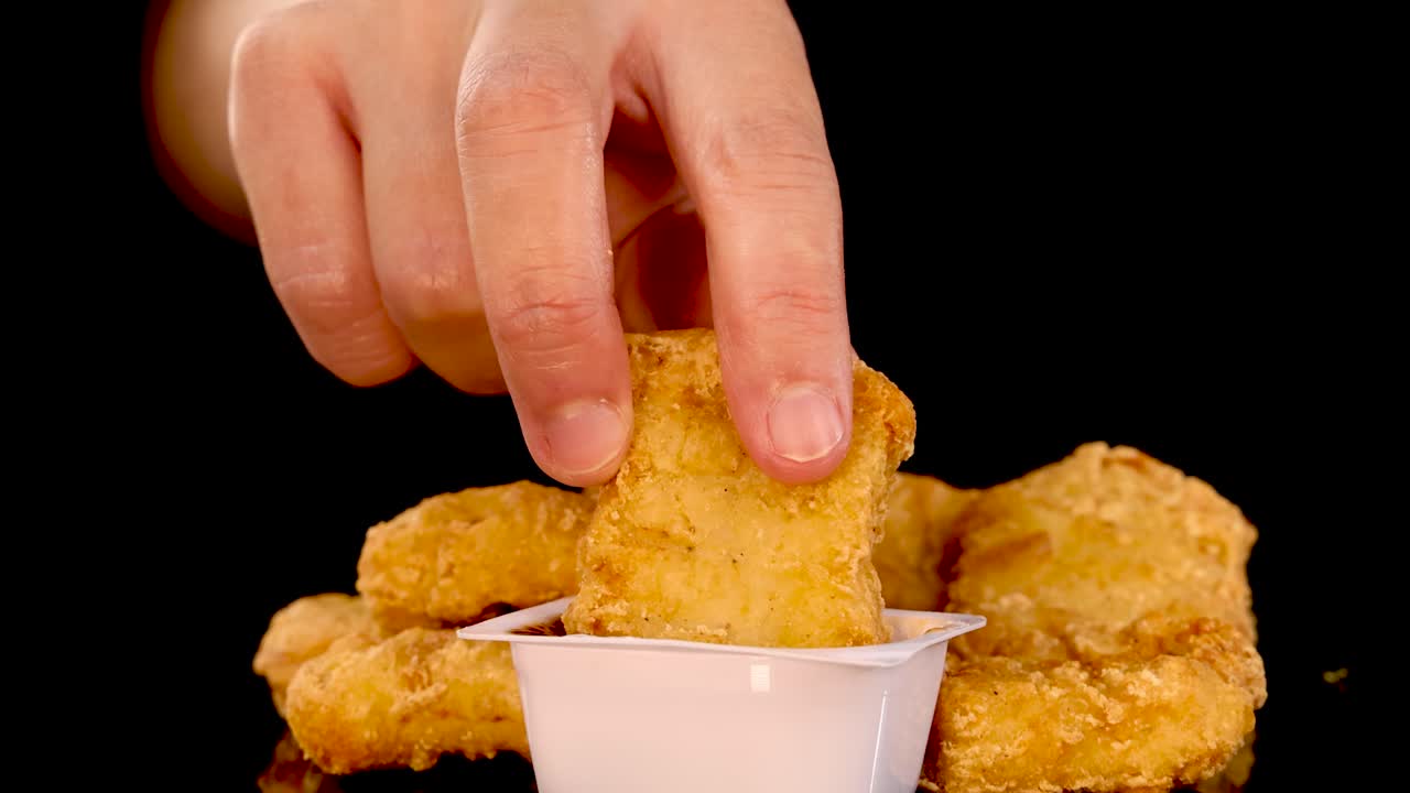 A close-up shot of a hand dipping a golden, crumbed chicken nugget into a container of barbecue sauce, with dramatic studio lighting and a black background