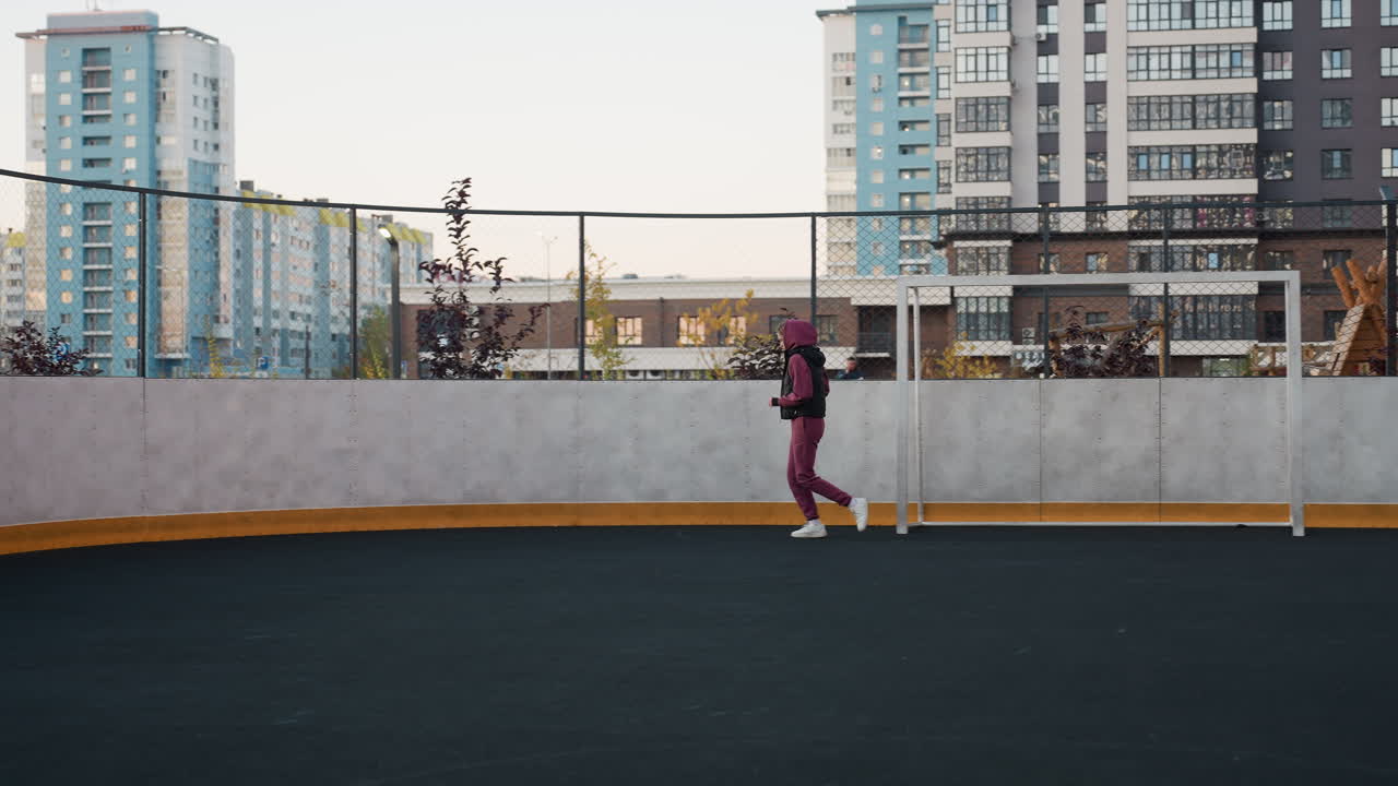 Female runner jogging around circular outdoor sports court next to white post under modern apartment building facades at dusk wearing hoodie vest joggers sneakers in steady motion