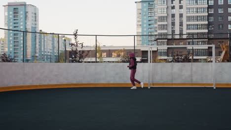 female runner jogging around circular outdoor sports court next to white post under modern apartment building facades at dusk wearing hoodie vest joggers sneakers in steady motion