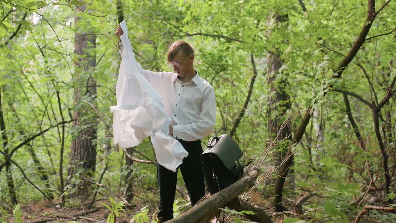 Young scientific researcher in white shirt taking out coat from black leather bag resting on tree branch in dense forest, surrounded by green leaves, trunks, branches, and natural sunlight