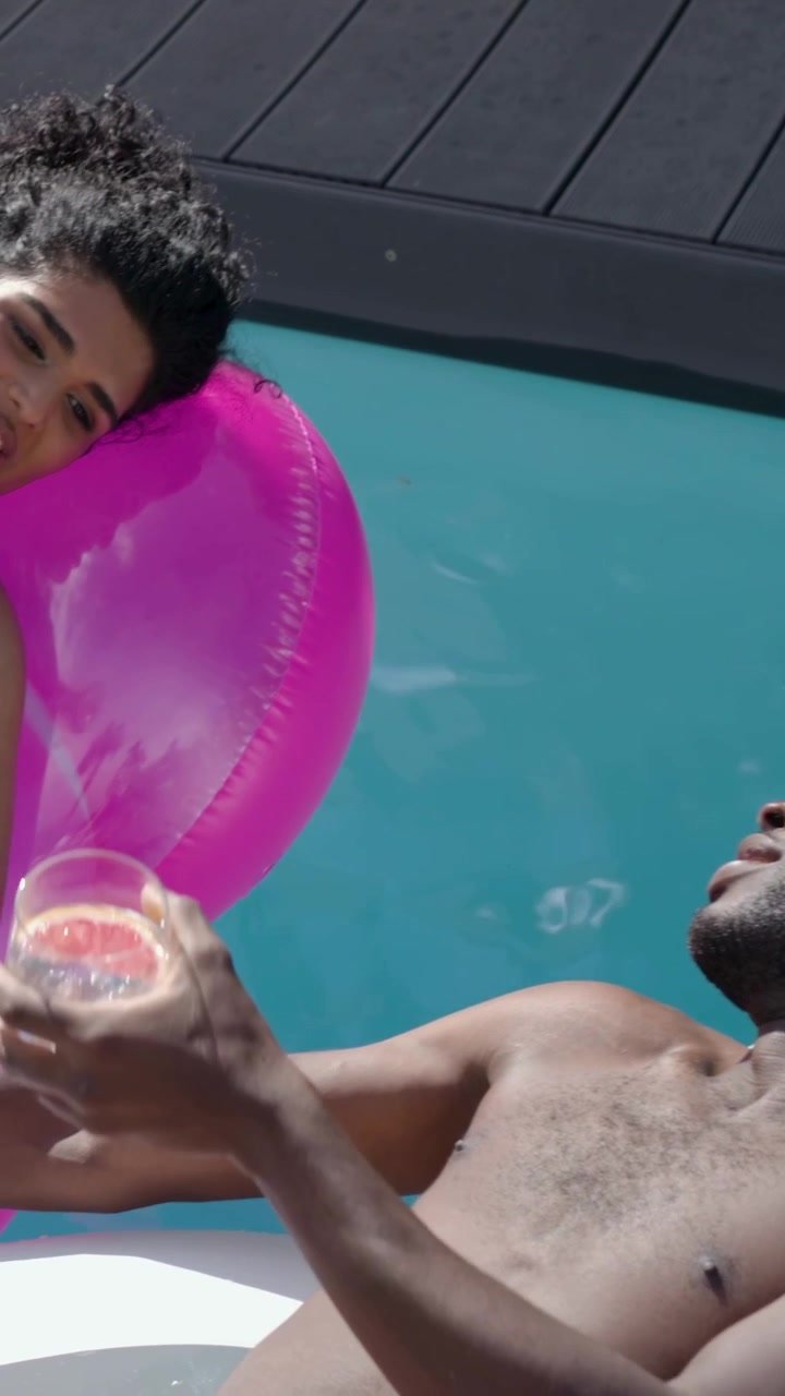 Vertical video: Woman leaning on pink ring, shirtless man extending arm offering pink glass at pool