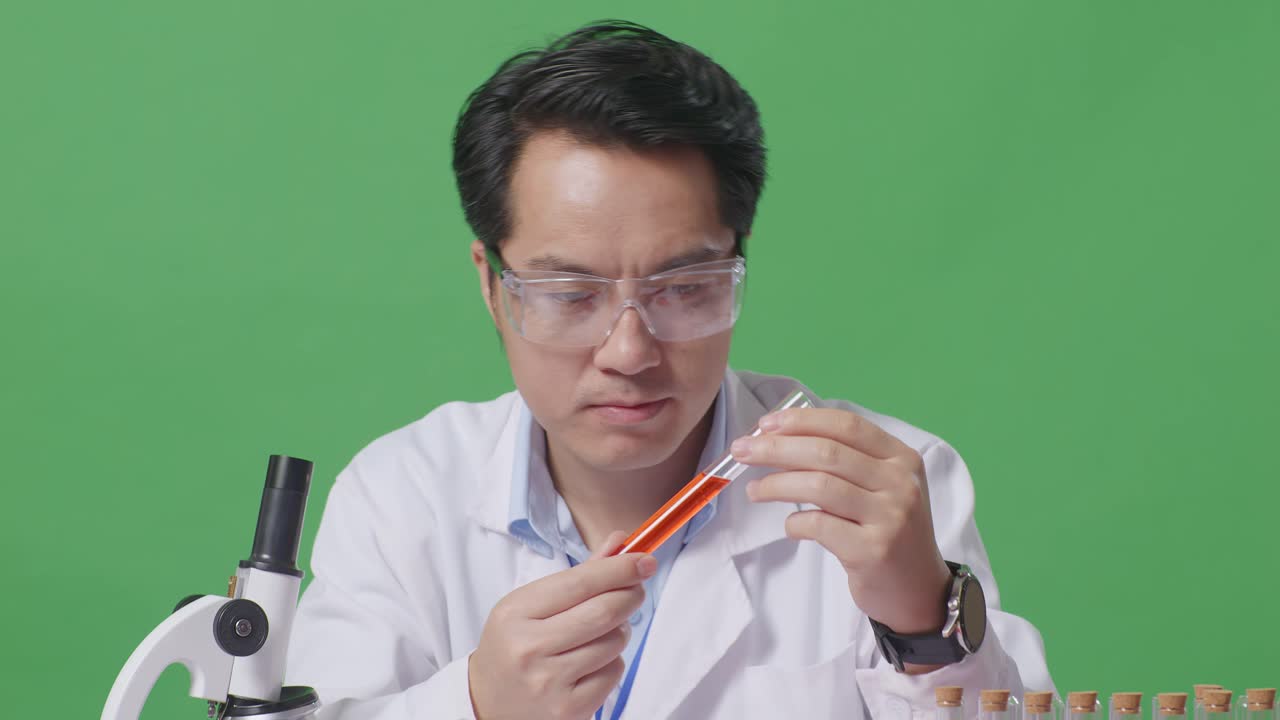 Close Up Of Asian Man Scientist Looking At The Orange Liquid In The Test Tube And Shaking Head On The Table With Microscope In The Green Screen Background Laboratory