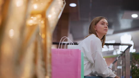 young woman seated on bench in shopping mall surrounded by vibrant decor and shopping bags, gazing thoughtfully at illuminated surroundings with blurred festive lights